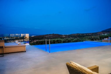 Gün batımında deniz ve tepelerin panoramik manzaralı sonsuz lüks havuz. Hasır mobilyalarla, yumuşak yastıklarla ve rulo havlularla dışarıda. Derin mavi gökyüzünün altında sakin bir ortam, mükemmel bir rahatlama.