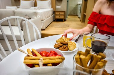 Kırmızı elbiseli bir kadının kahvaltı sahnesinde kruvasanlar, kurabiyeler, ekmekler, kiraz domatesleri, sade kahve, portakal suyu, ahşap zemin ve beyaz mobilyalar.