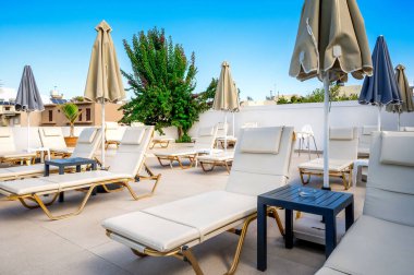 Beyaz şezlonglu, bej şemsiyeli ve modern açık hava manzaralı lüks güneş banyosu alanı. Açık mavi gökyüzü, yeşillik ve şehir binaları rahatlatıcı bir hava yaratıyor.