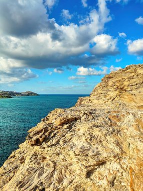 Engebeli kayalık kayalık sakin denizi, parlak mavi gökyüzünün altında dağınık bulutları görüyor. Kaya oluşumları, sakin derin mavi sulara zıt düşerek doğal güzellikle dramatik bir sahil manzarası yaratır.  
