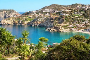 Turkuaz suları, kayalık uçurumları ve yeşil bitki örtüsüyle sersemletici Akdeniz Körfezi. Tepenin yamacında beyaz evleri olan küçük bir köy. Açık mavi gökyüzü huzurlu kıyı manzarasının güzelliğini vurguluyor