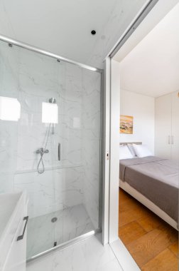 Cam duş muhafazalı parlak minimalist banyo, mermer fayanslar, rahat yatak odasını gri yatak odasına bağlayan sürgülü kapı ve temiz modern apartmanda beyaz gardırop..