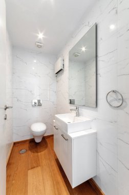 Ahşap zeminli modern minimalist banyo, duvara monte edilmiş tuvalet, beyaz mermer fayanslar, büyük ayna, lavabo ve krom detaylarıyla küçük beyaz gösteriş.