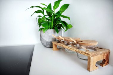 Ahşap kapaklı cam baharat kavanozları ve tahta kaşıklar yeşil bitki, modern beyaz mutfak detayı, pratik ve şık elementlerle doğal tonlarda..