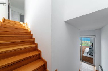 Modern minimalist evdeki parlak ahşap merdiven. Beyaz duvarları ve doğal ışığı var. Sürgülü cam kapıdan deniz manzaralı bir odaya açılıyor..