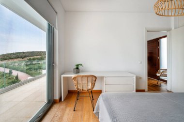 Minimalist yatak odası, doğal ahşap zeminler, beyaz masa ve sandalye, yeşil tepelerin manzarasına açılan büyük cam kapı ve sakin doğa manzarası..