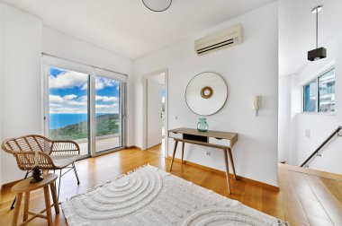 Sahil manzaralı parlak modern oda, minimalist dekor, doğal ahşap zemin, beyaz duvarlar, rattan sandalye, küçük masa, büyük pencereler, sakin ve havadar iç mekan..