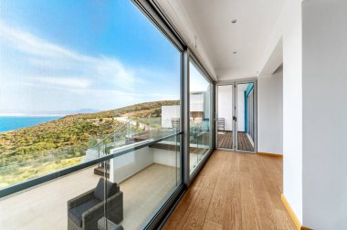 Büyük cam kapıları ve dağ manzaralı güneş ışığı koridoru. Ahşap zeminli ve parlak çizgileri olan modern balkon alanı iç mekanla açık hava bağlantısı oluşturur..