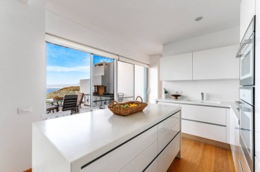 Ada tezgahlı parlak beyaz mutfak, modern dolaplar, meyve sepeti ve veranda kapıları okyanus manzarası ve açık oturma alanına açılıyor..