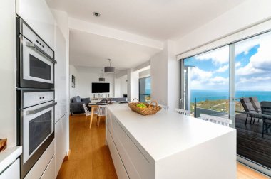 Ada, meyve sepeti, geniş pencereli panoramik okyanus manzaralı, açık planlı yemek ve yaşam alanına bağlı modern beyaz mutfak..