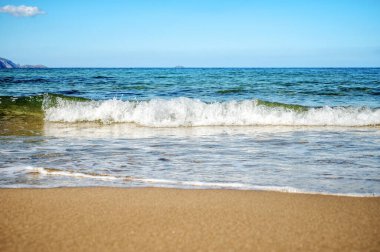 Yumuşak okyanus dalgaları açık mavi gökyüzünün altında temiz kumlu kumsala yuvarlanıyor. Huzurlu deniz manzarası, parlak gün ışığında yansıma ve ufuk çizgisiyle yükselen suyu yakalar..