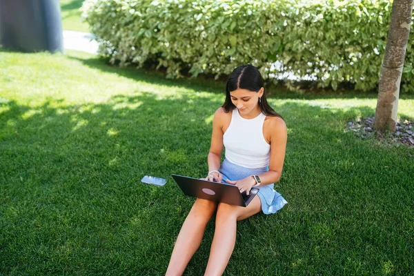 The concept of remote work. A young girl works with a laptop in the fresh air in the park, sitting on the lawn.