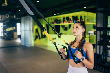 Spor kıyafetleri içinde mükemmel vücutlu genç atletik bir kadın spor salonunda trx fitness kayışlarıyla egzersiz yapıyor.