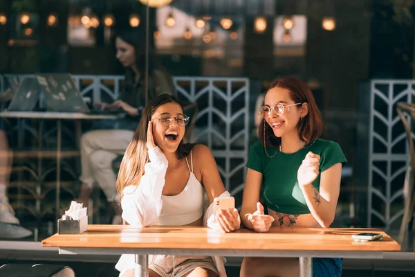 屋外のカフェに座っている間に携帯電話を使用して2人の興奮した若い女の子 — ストック写真
