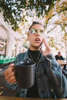 Genç bir kadın akıllı telefondan arıyor ve şehirdeki kafe terasında içki içiyor.