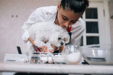 Mutfakta gülümseyen genç bir kadın elinde sevimli beyaz bir Malta köpeği tutuyor.