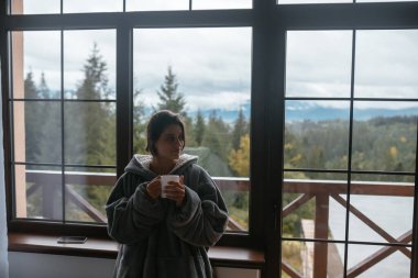 Pencerenin yanında duran kadın elinde bardak tutuyor.