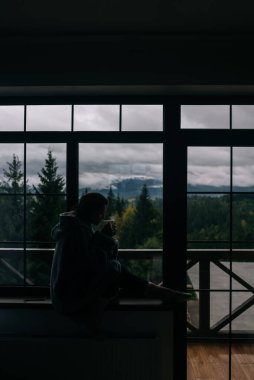 Pencerenin kenarında elinde kupayla oturmuş dağlara ve yeşil doğaya bakan bir kadının silueti.