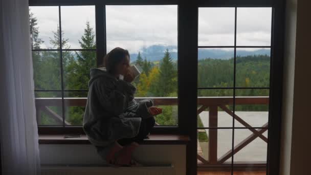 窓辺に座って山と緑の自然を眺めながら熱いコーヒーを飲んでいる美しい女性の肖像 — ストック動画