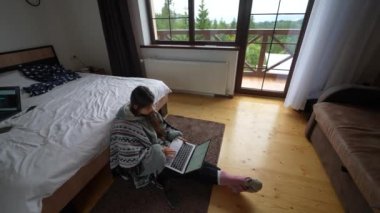 Diz üstü bilgisayarı olan meşgul genç bir kadın çift kişilik yatağın yanında oturuyor ve ağ kuruyor ya da çeşitli web sitelerinde boş zaman geçiriyor.