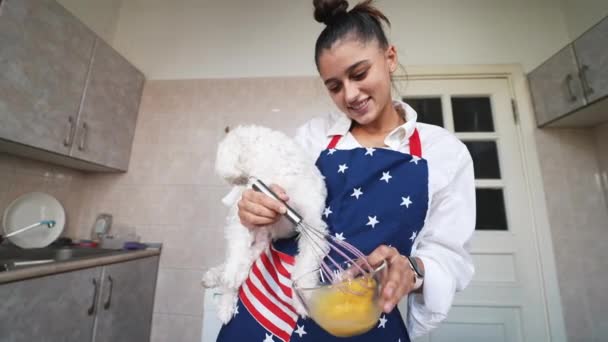 笑顔の若い女がキッチンで卵をささやきながら可愛い白いマルチ犬を抱えています — ストック動画