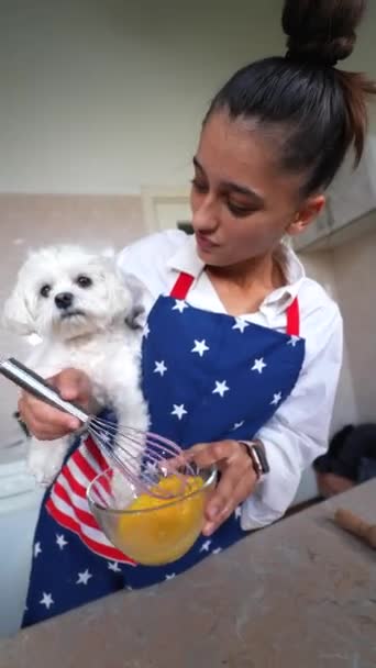 笑顔の若い女がキッチンで卵をささやきながら可愛い白いマルチ犬を抱えています — ストック動画