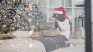 Pencereden bakan genç bir kadın, elinde Noel ağacının yanında bir hediye tutuyor.