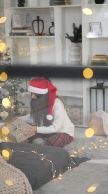 Pencereden bakan genç bir kadın, elinde Noel ağacının yanında bir hediye tutuyor.