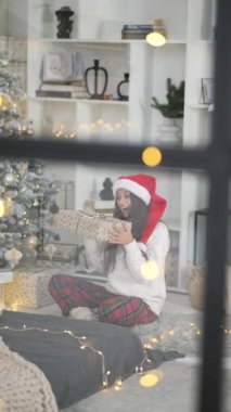 Pencereden bakan genç bir kadın, elinde Noel ağacının yanında bir hediye tutuyor.
