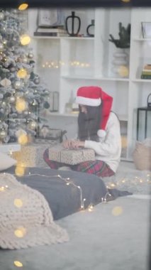 Pencereden bakan genç bir kadın, elinde Noel ağacının yanında bir hediye tutuyor.