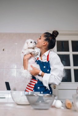 Mutfakta gülümseyen genç bir kadın elinde sevimli beyaz bir Malta köpeği tutuyor.