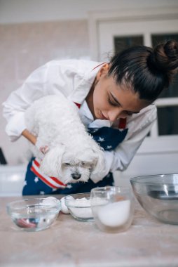 Mutfakta gülümseyen genç bir kadın elinde sevimli beyaz bir Malta köpeği tutuyor.