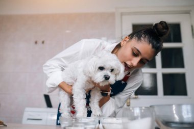 Mutfakta gülümseyen genç bir kadın elinde sevimli beyaz bir Malta köpeği tutuyor.
