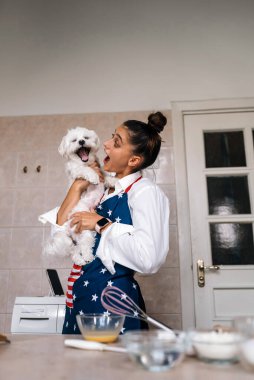 Mutfakta gülümseyen genç bir kadın elinde sevimli beyaz bir Malta köpeği tutuyor.