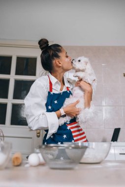 Mutfakta gülümseyen genç bir kadın tatlı beyaz Malta köpeğini öpüyor.