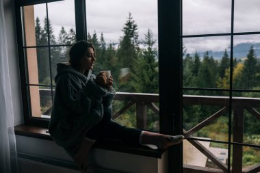 Pencerenin kenarında elinde kupayla oturmuş dağlara ve yeşil doğaya bakan bir kadının silueti.