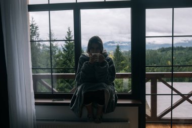 Pencerenin kenarında elinde kupayla oturmuş dağlara ve yeşil doğaya bakan bir kadının silueti.
