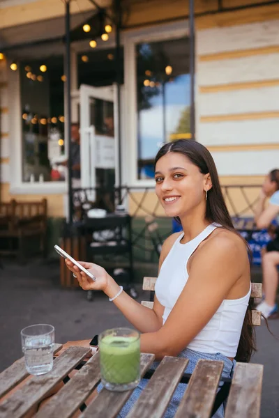 Smiling woman holding phone outdoors in cafe. A beautiful model looks at the camera