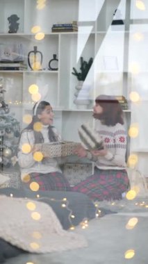 Pencereden bakan iki kız arkadaş, Noel ağacının yanında oturmuş hediyelerini değiş tokuş ediyorlar.