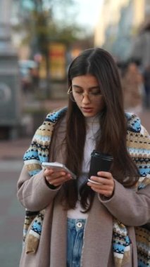 Kalabalık şehir caddesinde elinde kahve fincanı ve cep telefonuyla yürüyen güzel bir kadın.