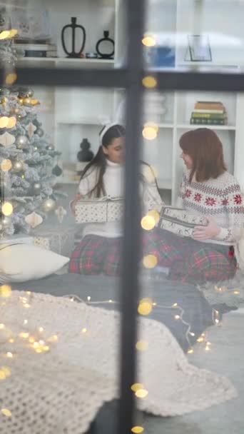 Смотря Окно Две Девушки Обмениваются Рождественскими Подарками Сидя Рядом Елкой — стоковое видео