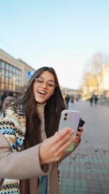 Mutlu gülümseyen genç bir kadın şehirde video görüşmesi yapıyor. İnternet günlüğü için selfie ya da video çeken şık bayan gezgin.