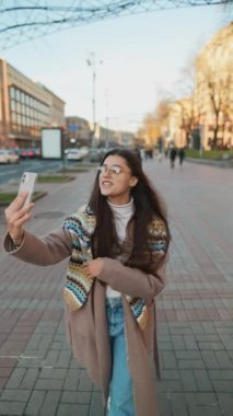 Mutlu gülümseyen genç bir kadın şehirde video görüşmesi yapıyor. İnternet günlüğü için selfie ya da video çeken şık bayan gezgin.