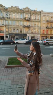 Mutlu gülümseyen genç bir kadın şehirde video görüşmesi yapıyor. İnternet günlüğü için selfie ya da video çeken şık bayan gezgin.