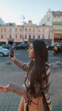 Mutlu gülümseyen genç bir kadın şehirde video görüşmesi yapıyor. İnternet günlüğü için selfie ya da video çeken şık bayan gezgin.