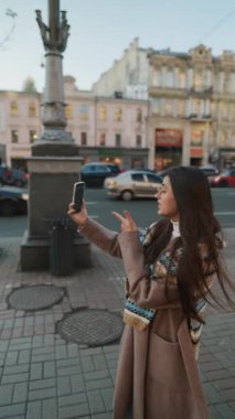 Mutlu gülümseyen genç bir kadın şehirde video görüşmesi yapıyor. İnternet günlüğü için selfie ya da video çeken şık bayan gezgin.