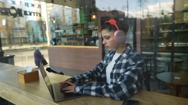 ヘッドフォンの美しい女の子がカフェの窓の後ろのノートパソコンに入力しています 高品質のフルHd映像 — ストック動画