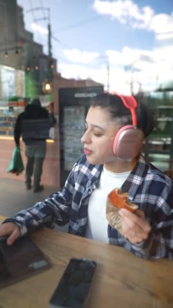戴着耳机的漂亮姑娘在咖啡馆里用笔记本电脑吃午饭 听音乐 优质Fullhd影片 — 图库视频影像
