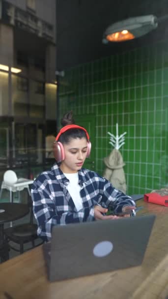 カフェの窓の後ろのノートパソコンに入力するヘッドフォンの美しい女の子 高品質のフルHd映像 — ストック動画