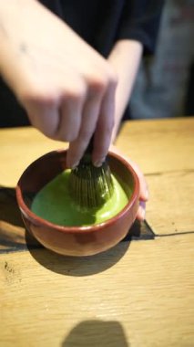 Ahşap masa arkasında beyaz kasede yeşil çay yapma süreci. Kahverengi masada fincanda kibrit yeşil çay. Kadın, ahşap kaşıkla kibrit kutusuna organik yeşil çay tozu döküyor.. 